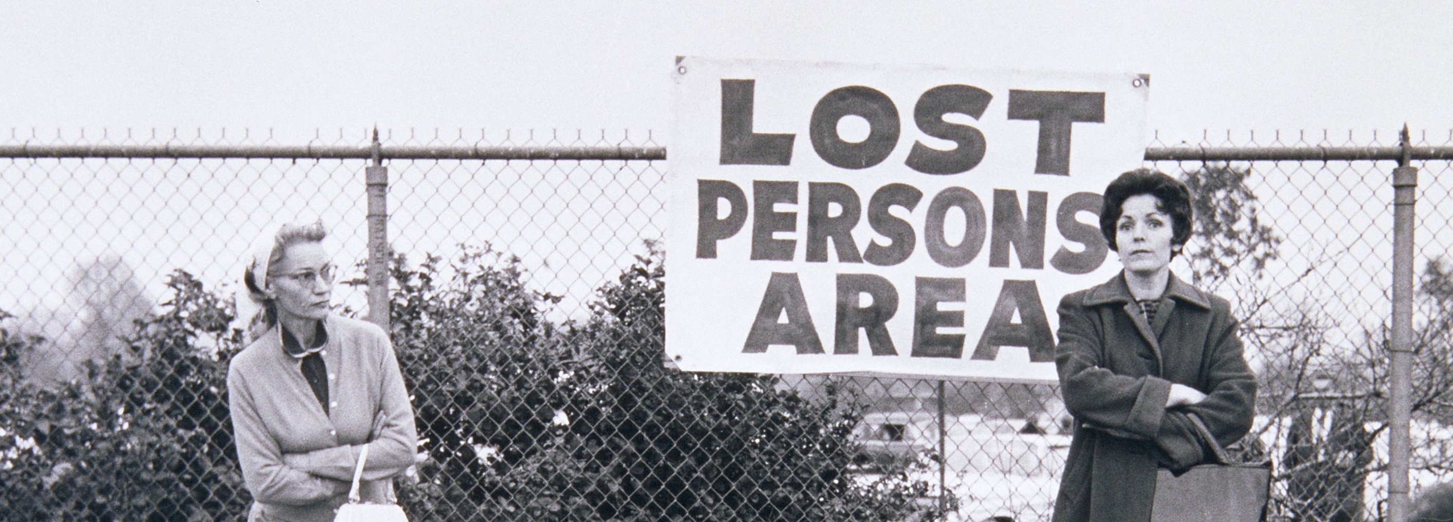 a black and white photo of several women standing beside a chainlink fence and a handmade sign which reads "Lost Persons Area"